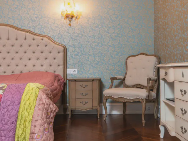 Łóżko z różowym pikowanym zagłówkiem, błękitna tapeta w ornament, rzeźbiony fotel, komoda i kinkiet