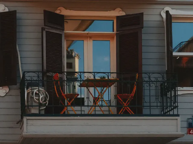 Balkon domu z czarną kutą balustradą, drewnianą podłogą, pomarańczowymi składanymi krzesłami i stolikiem przy oknie z okiennicami