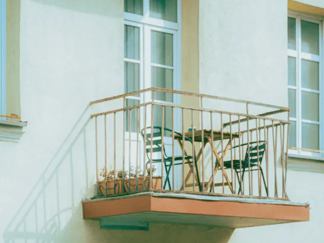 Niewielki balkon w pastelowej kamienicy z metalową balustradą, zielonymi krzesłami, stolikiem i skrzynkami z kwiatami