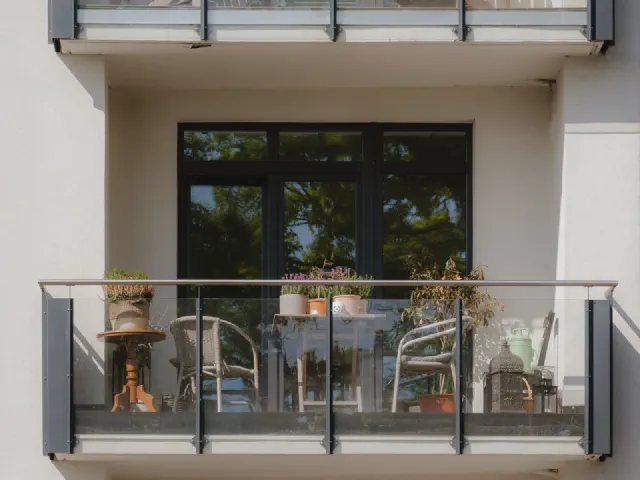 Balkon w bloku ze szklaną balustradą, widziany z zewnątrz – krzesła, stolik i rośliny w donicach przy przeszklonych drzwiach