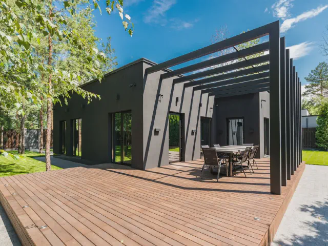 Duży drewniany taras przy nowoczesnym czarnym domu, nad częścią tarasu czarna pergola z belek, w głębi stół z krzesłami