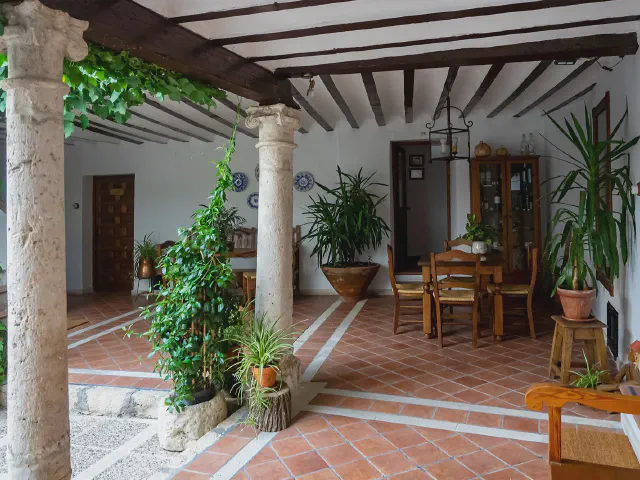 Patio w stylu hiszpańskim z kolumnami, drewnianym stropem z belek, podłogą z terakoty, drewnianym stołem i dużą ilością roślin w donicach