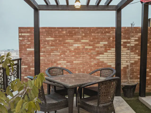 Drewniana pergola nad stołem z rattanowymi krzesłami przy ceglanym murze, obok rośliny w donicach