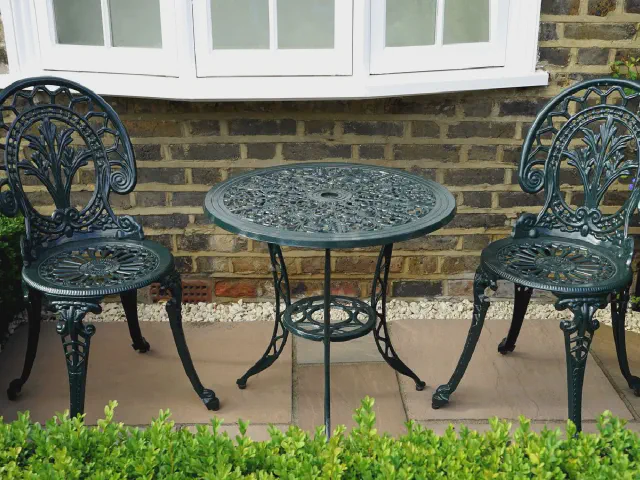 Czarny żeliwny stolik i dwa krzesła na tarasie z płyt przy ceglanej ścianie domu, przed nimi niski żywopłot