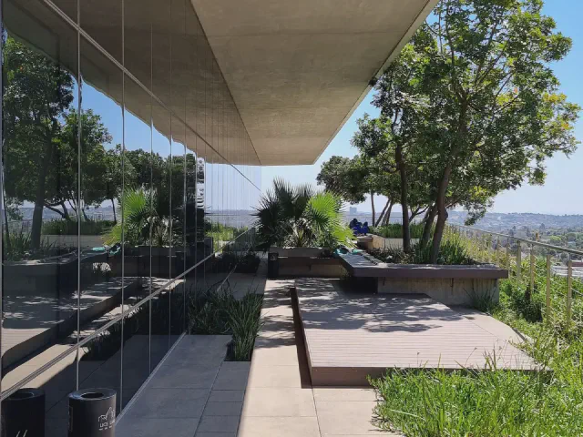 Taras z płyt betonowych pod betonowym nawisem budynku, ze szklaną balustradą, rabatami i widokiem na miasto